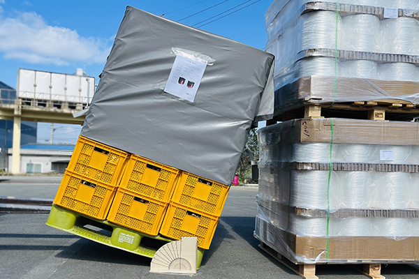 Grey pallet cover over yellow crates tilting precariously on a green pallet, demonstrating stability or a tilt test outdoors